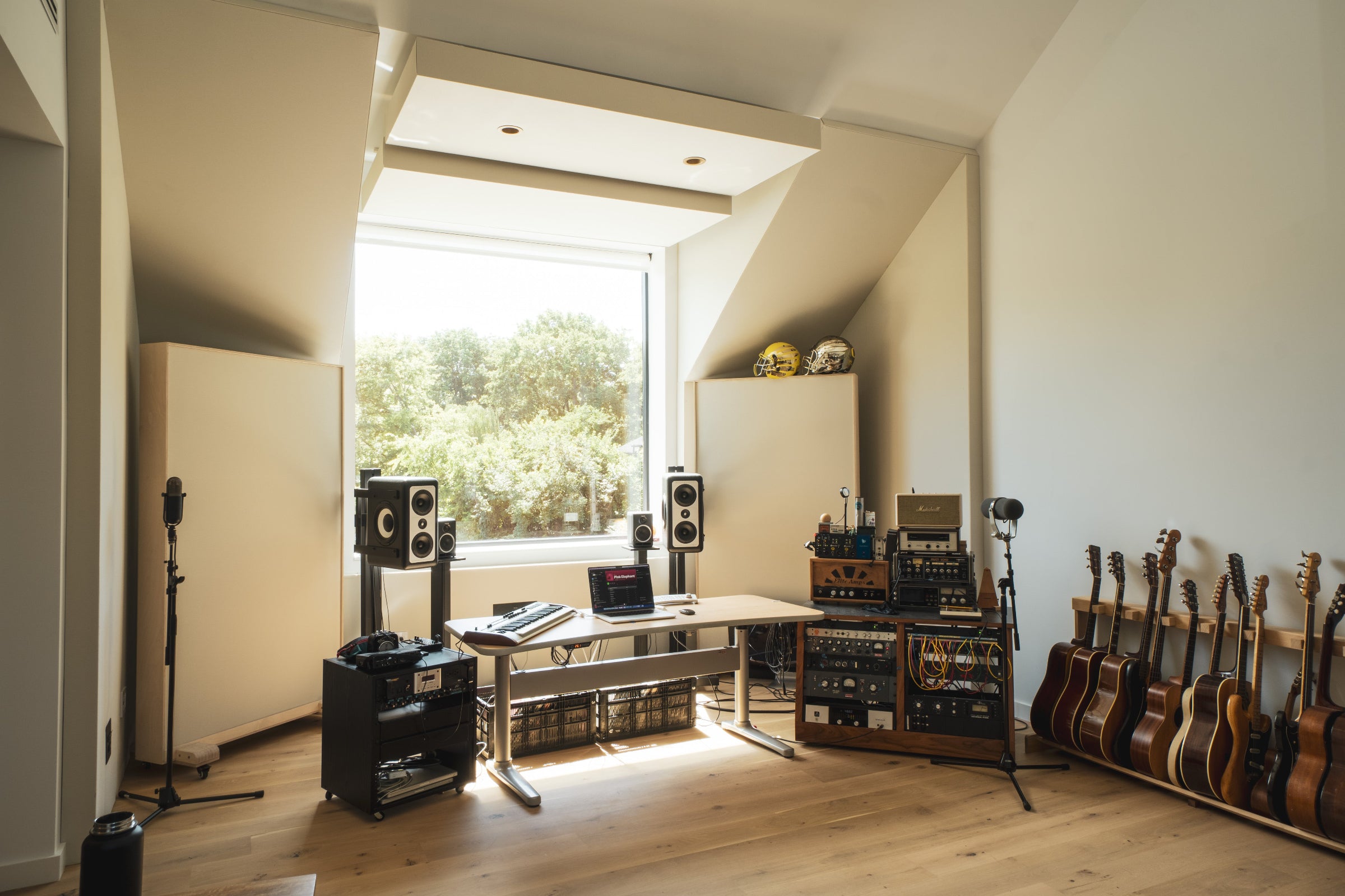 Home music studio with acoustic panels, guitars along one wall, recording equipment on a desk, and large window providing natural light.