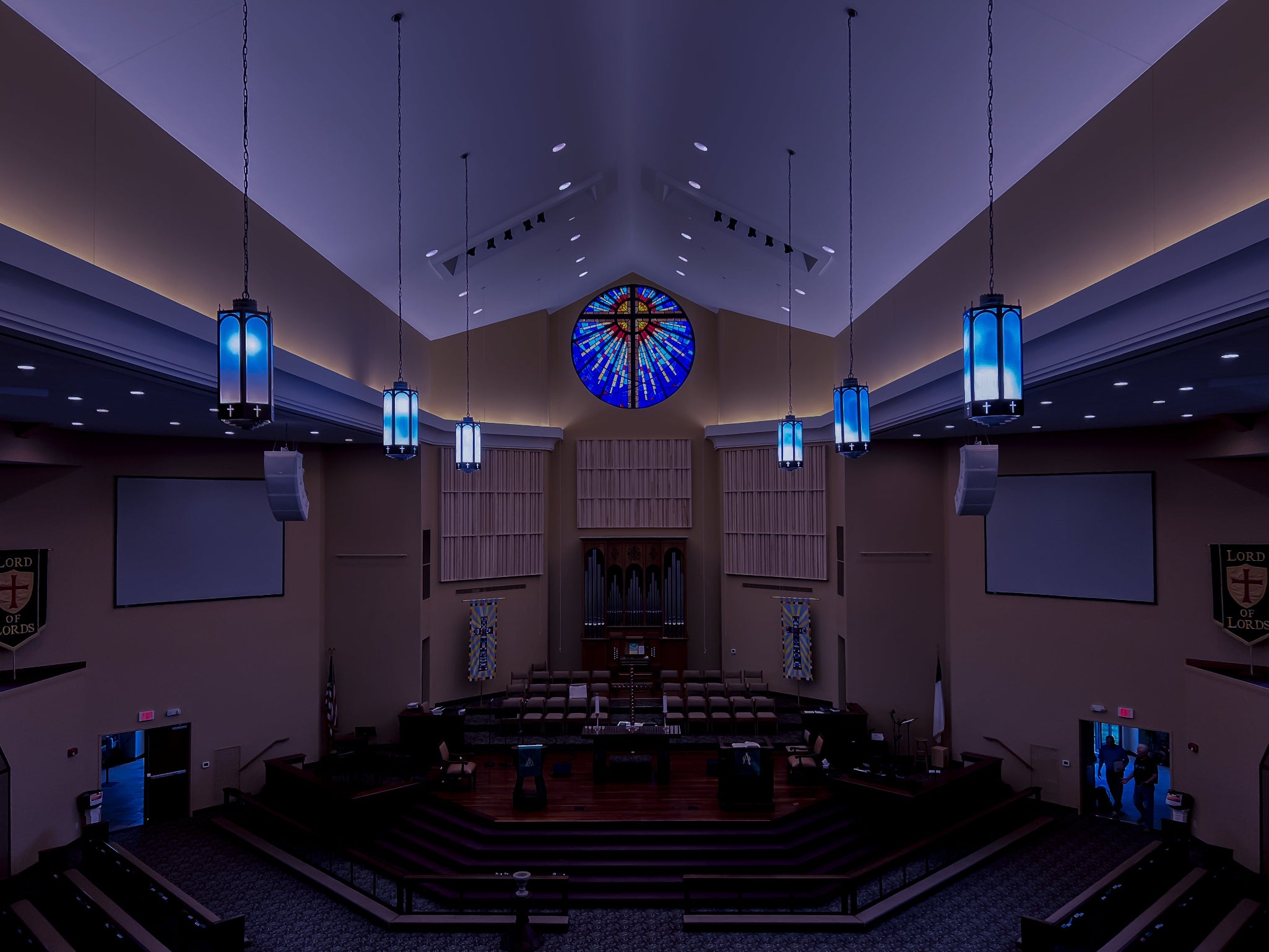 Interior of a church with a large stained glass window and blue pendant lights.